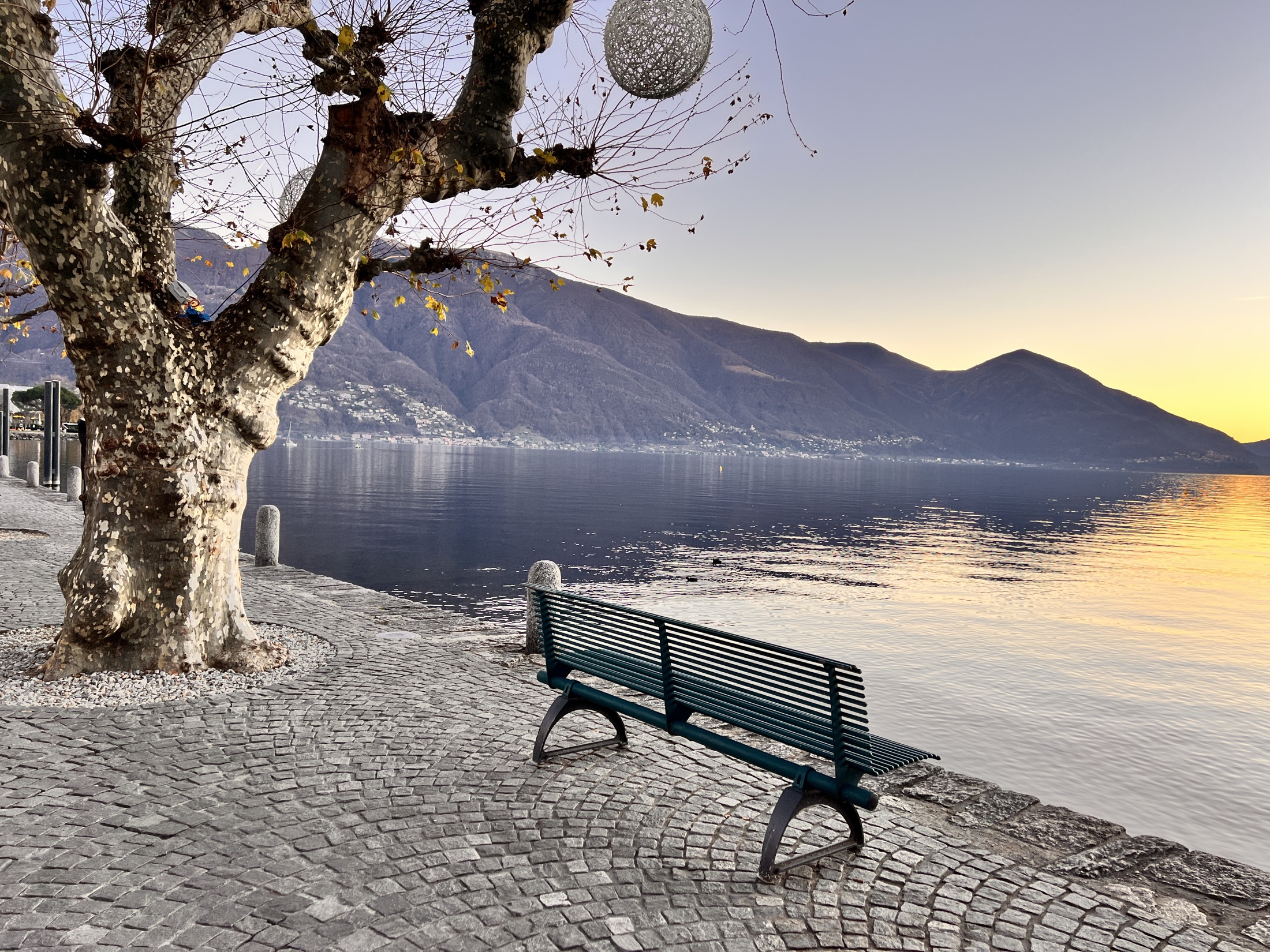 Empty green bench on a cobbled lakeside promenade at golden hour, calm blue water and mountains beyond